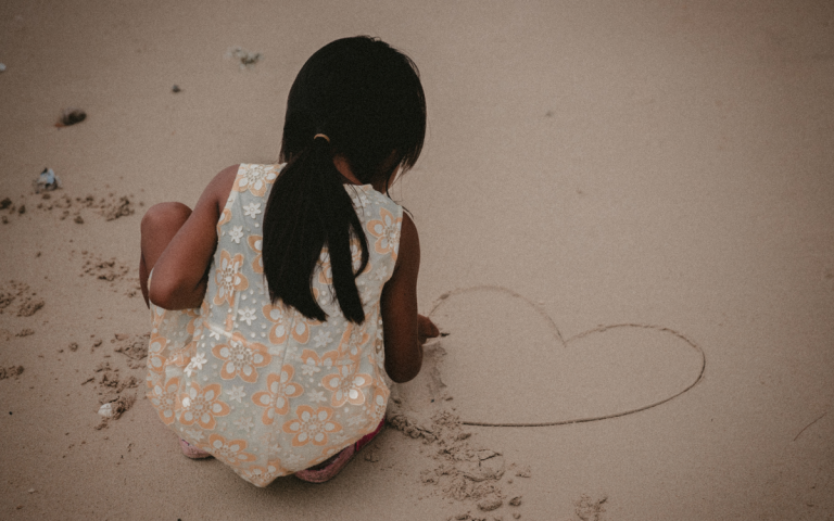 Jugando con arena.Una niña con un vestido de flores, se encuentra agachada en la arena de una playa realizando una silueta de un corazón, con un objeto que se encontro en la playa. foto por Jakob Owens, stocksnap.io